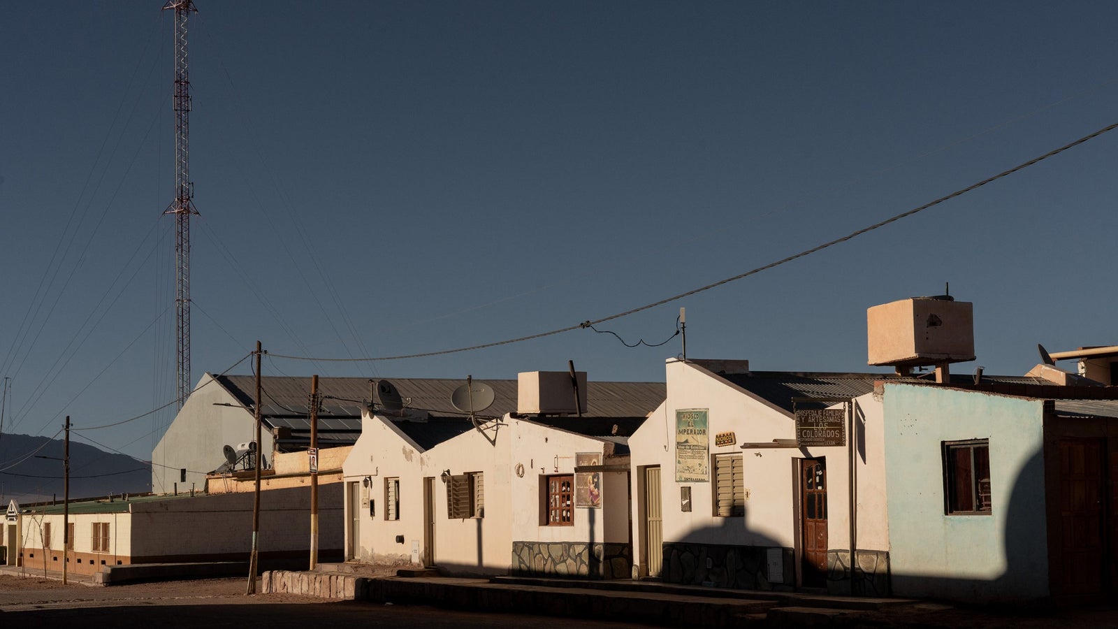 A street scene featuring modest, white-walled buildings with varying architectural details, including windows with wooden shutters and signs, set against a clear blue sky. A communication tower rises in the background, alongside power lines and satellite dishes on the rooftops.