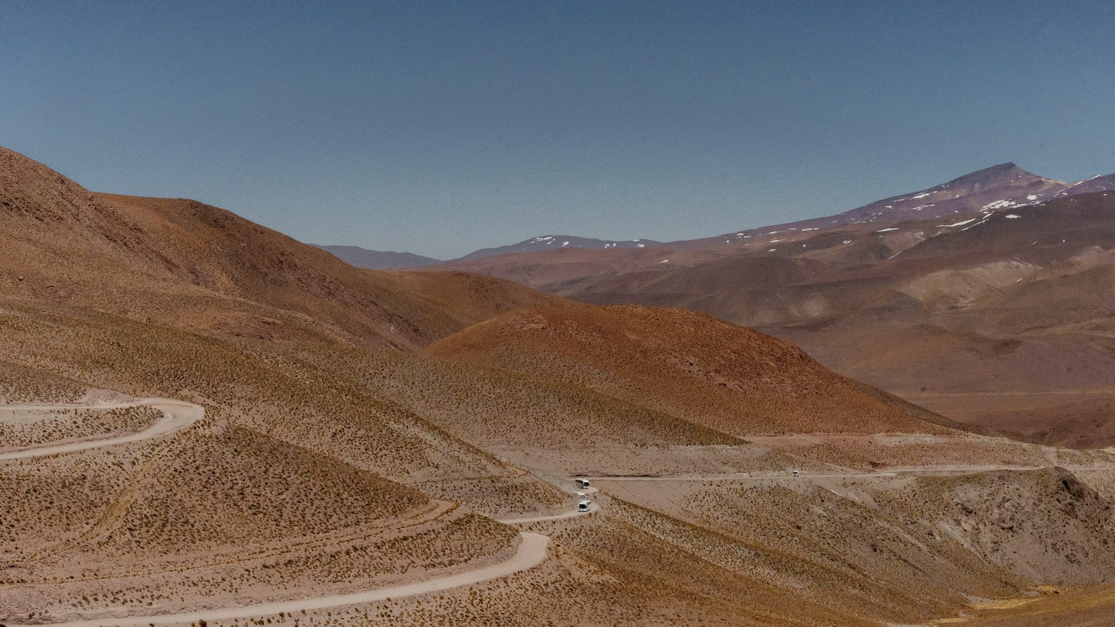 A winding dirt road traverses a barren, hilly landscape under a clear blue sky, with mountains in the background and sparse vegetation along the road.