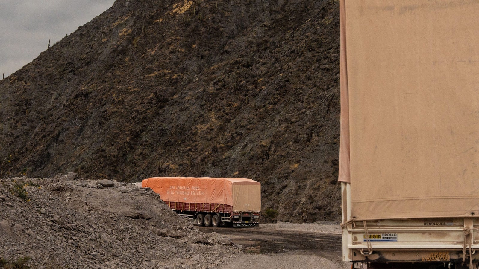 Two trucks on a gravel road winding through a mountainous area, with one truck parked and covered with a large orange tarp, while the other truck is partially visible, showing its rear lights against a rocky landscape.
