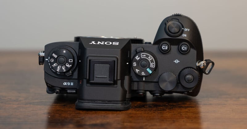 Top view of a black Sony digital camera showing various control dials and buttons, including the mode dial, exposure compensation dial, shutter button, and custom buttons, placed on a wooden surface.