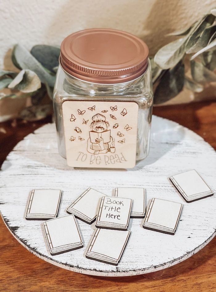 a clear class jar with a wooden placque on the front. The plaque has butterflies and a person reading a book on it anf text that reads To Be Read. In front of the jar are multiple small white wood tiles in the shape of books