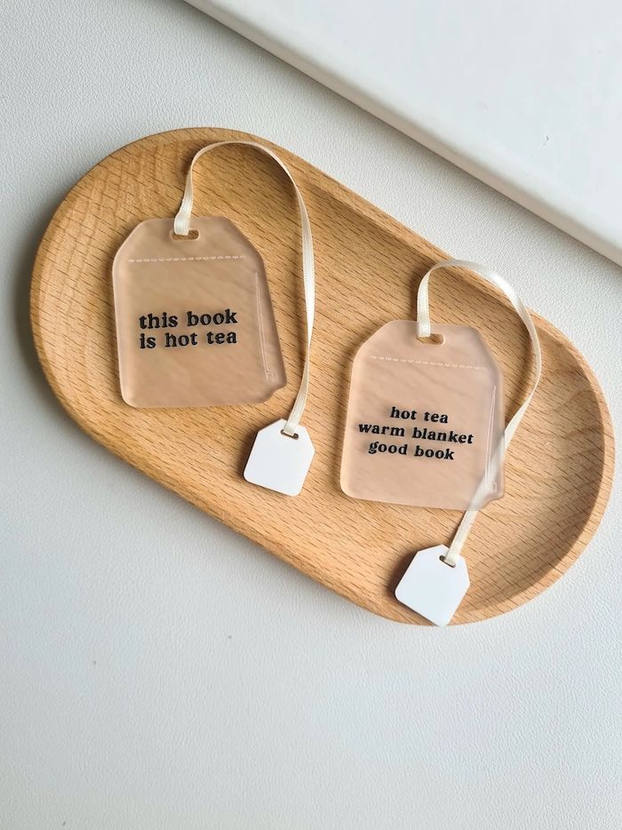 two clear acrylic bookmarks in the shape of tea bags resting on a wooden tray. both bookmarks have ribbons in them to look like strings on a tea bag and black text on them. One says This Book is Hot Tea, the other Hot Tea Warm Blanket Good Book