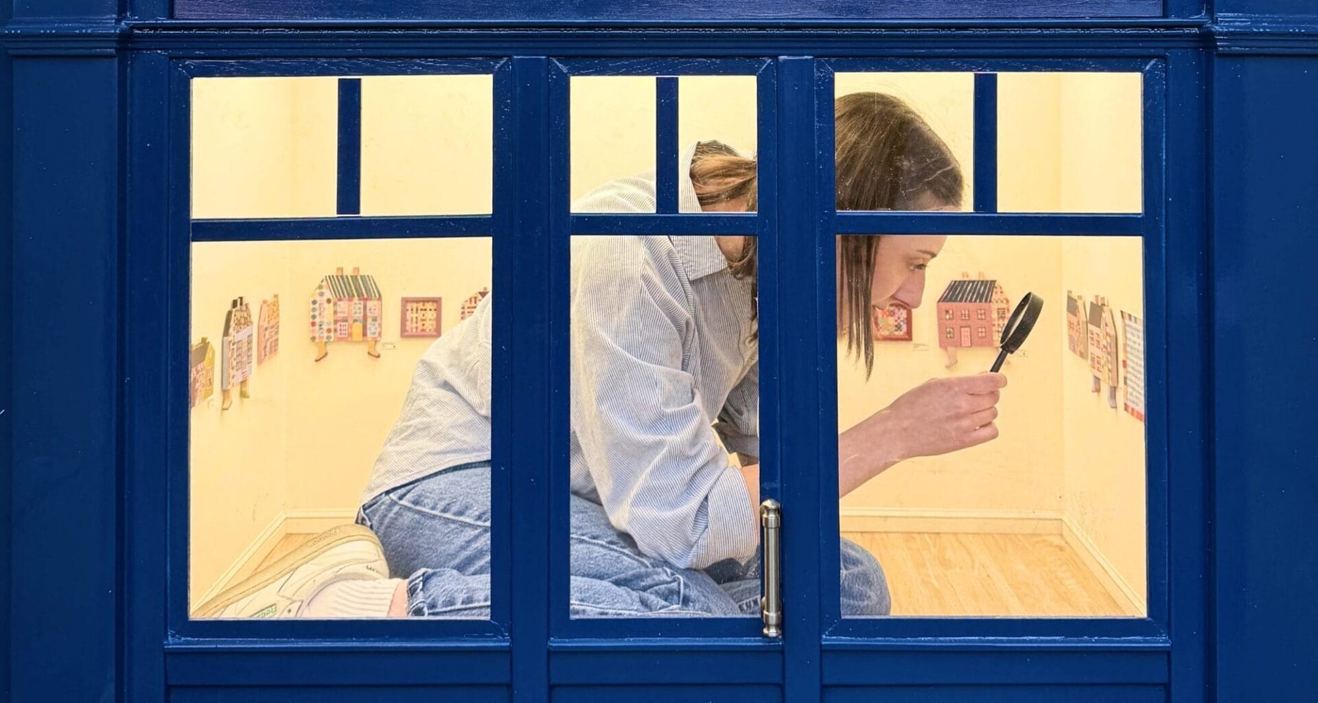 a person kneels inside of a 1/6-scale gallery, holding a magnifying glass up to a painting