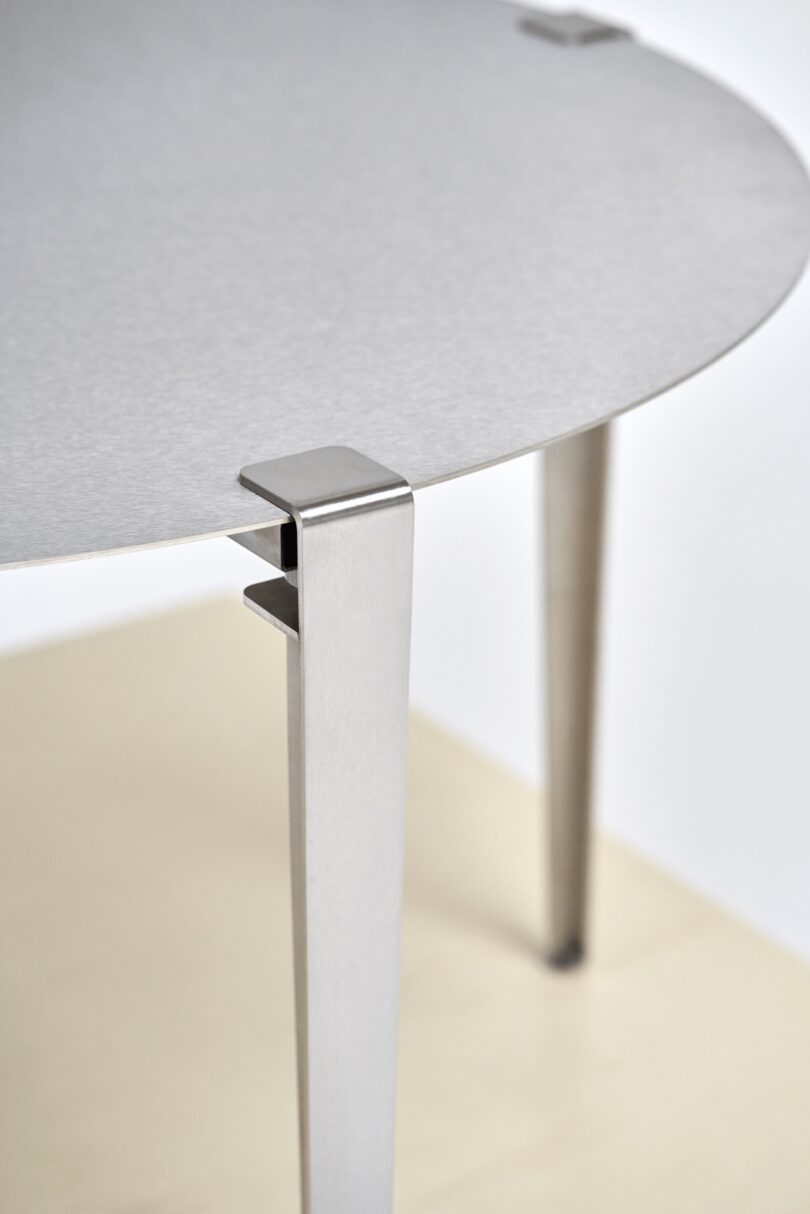 Close-up of a round, light gray metal table with square-edged legs and visible metal corner brackets, standing on tiptoe-like legs atop a light-colored surface