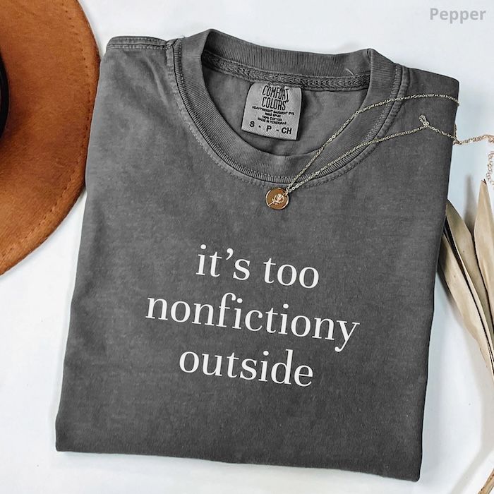 dark grey folded tshirt with white text on the front that reads "it's too nonfictiony outside"
