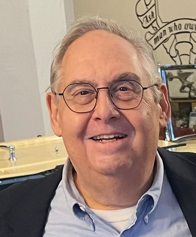 A smiling older man with glasses, short gray hair, and a navy blazer stands indoors with a drum set and a wall mural in the background.