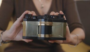 A person in a brown top holds a vintage-style camera with gold and black detailing, displaying it toward the viewer. The camera has a unique, boxy design and the person's hands frame it on each side.