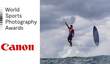 A surfer wearing a pink shirt appears to be mid-air above the ocean, with his surfboard flying nearby. The sky is cloudy. The left side features the text "World Sports Photography Awards" and the Canon logo.