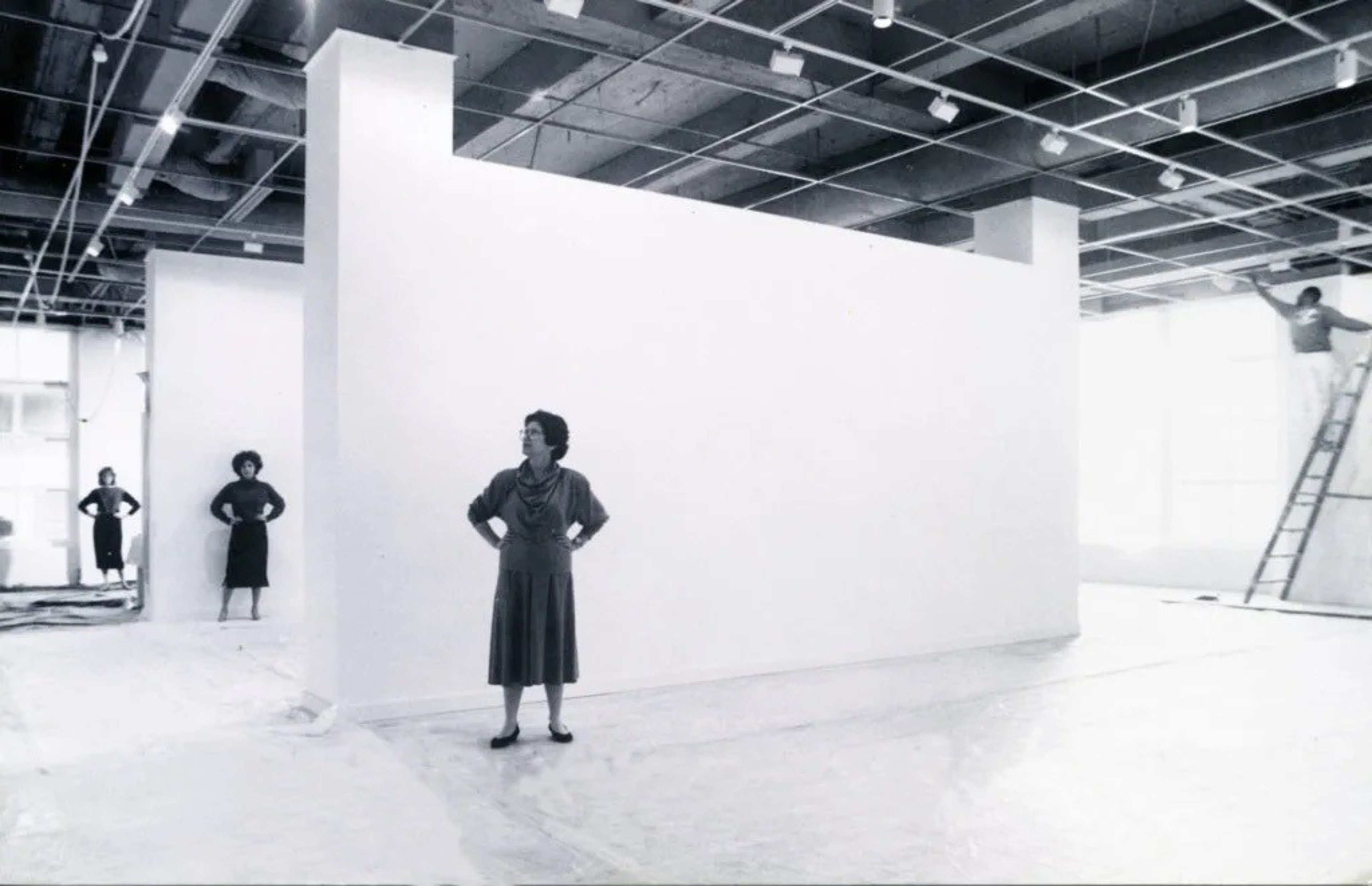 Three women stand with hands on hips spaced apart inside a large, empty gallery with white walls, while a person climbs a ladder on the right.