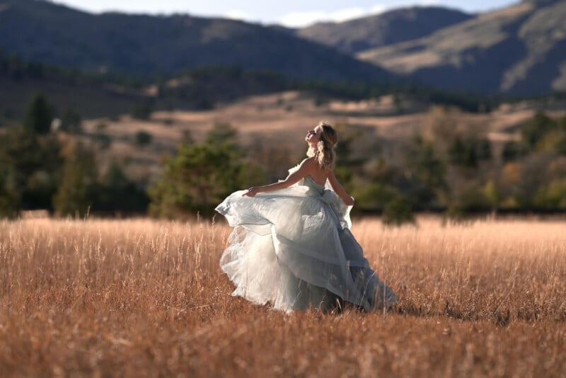 A woman in a flowing, light blue gown twirls in a golden field with mountains and trees in the background under a partly cloudy sky.