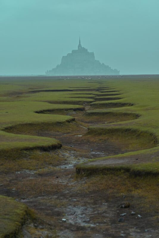 A misty landscape with cracked, grassy ground leading toward a distant, island abbey silhouetted against a pale blue sky. The scene feels serene and mysterious.