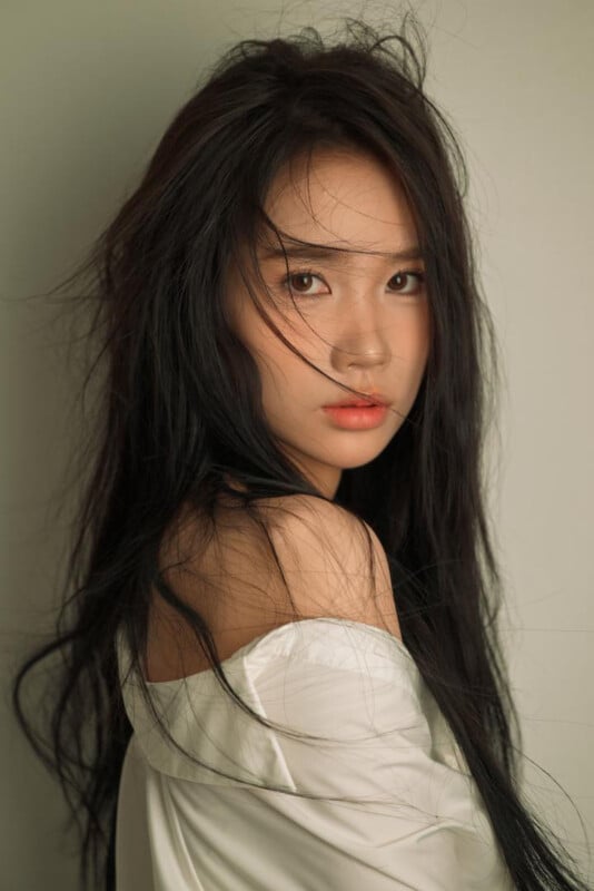 A young woman with long, dark hair and natural makeup looks over her shoulder. She wears a white off-shoulder top and gazes softly at the camera against a neutral background.