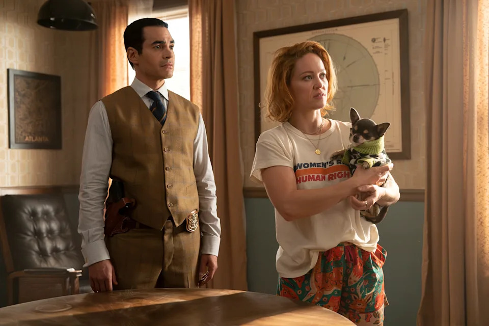 A man in a formal vest and tie stands with a woman in casual attire holding a small dog. They are in a room with vintage decor