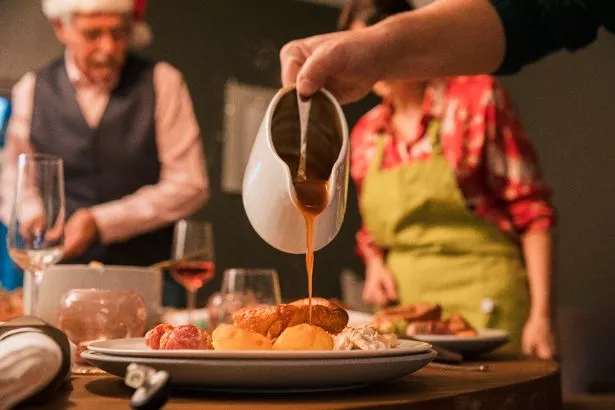 Gravy being poured from a jug onto a plate of Christmas lunch. A woman is wearing an apron out of focus in the background and there are more plates and glasses on the table ready for the family to sit down.