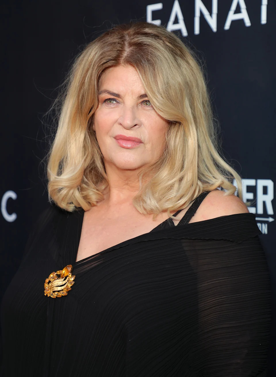 Person on red carpet wearing an elegant off-shoulder black dress with a gold brooch, posing in front of a backdrop with text