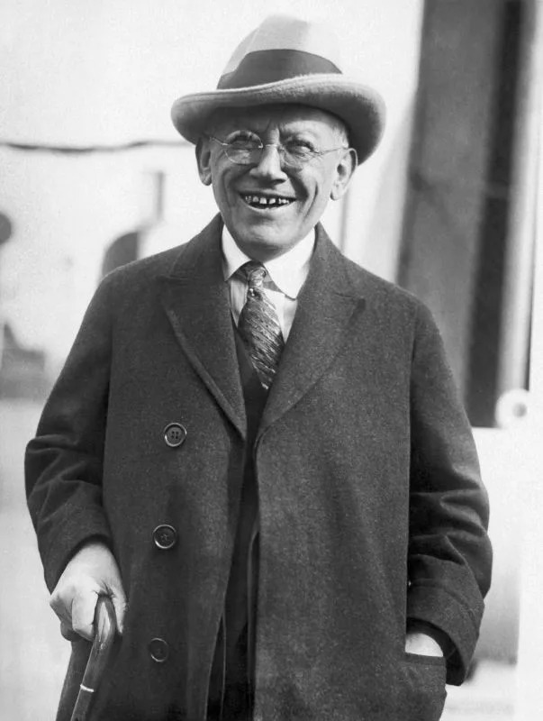 Smiling Laemmle in a suit, tie, and coat with glasses and a hat, holding an umbrella