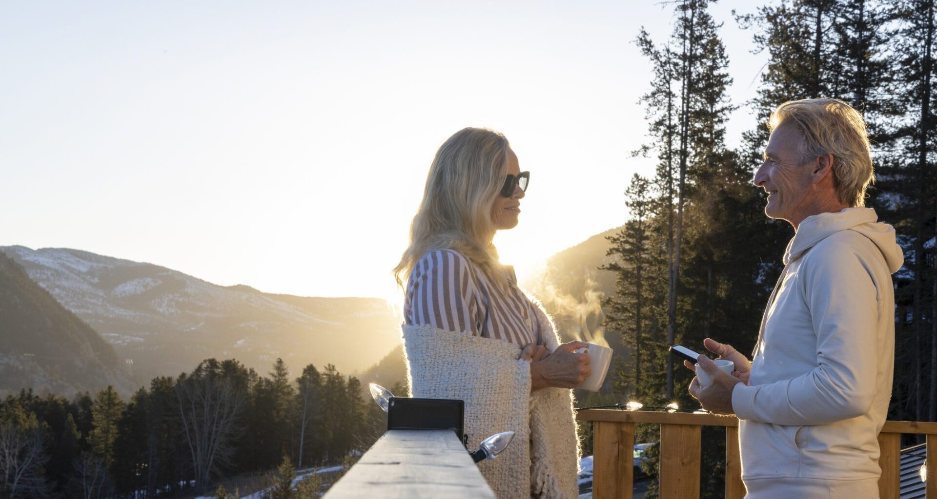 Mature couple chat on chalet deck at sunrise in winter, with phone and hot drink, Canadian Rockies.