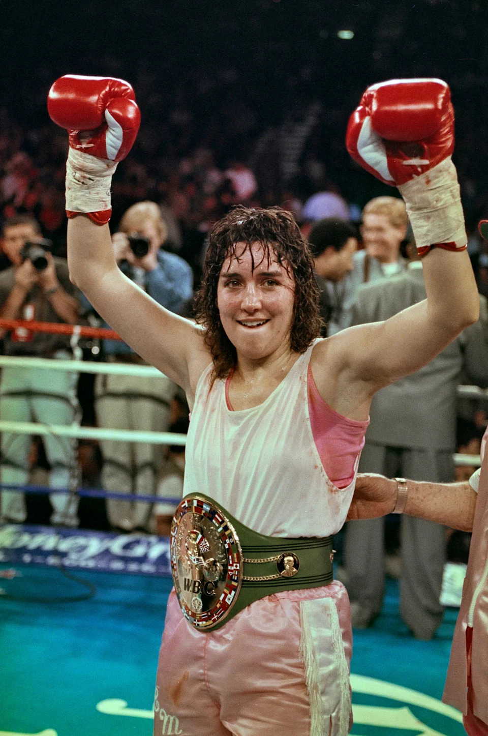 A victorious boxer with gloves raised, wearing a championship belt, smiles in the boxing ring. Photographers are visible in the background