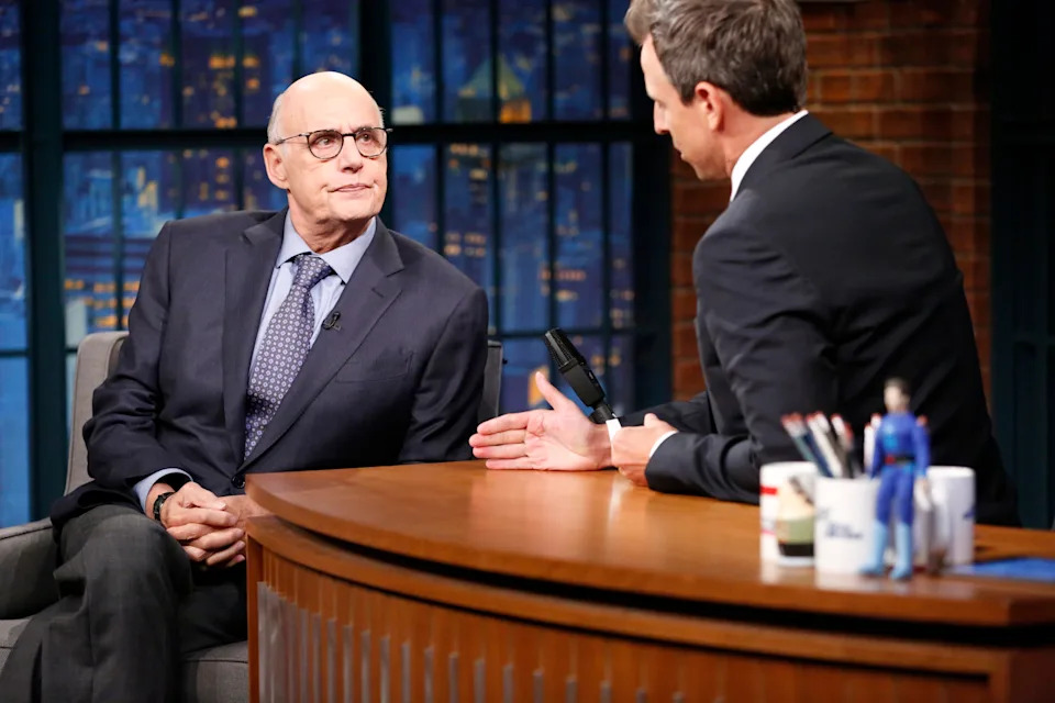 Two people in formal suits conversing on a late-night talk show set, seated at a desk with mugs and a small figure