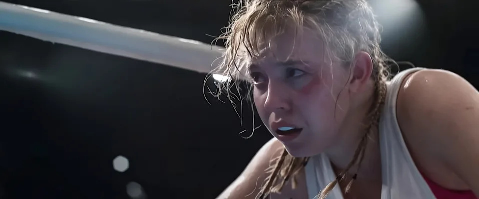 A woman in a boxing ring looks exhausted and determined, hair tousled, wearing a tank top and mouthguard, likely from a dramatic scene in a film
