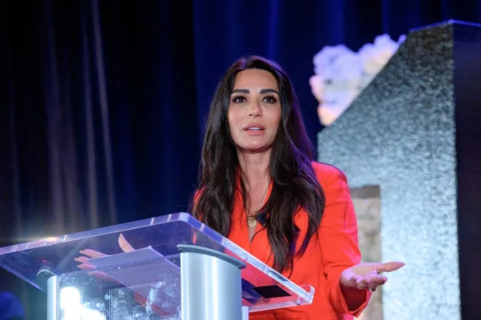 A woman speaks at a podium, gesturing with her hands. She wears a stylish blazer