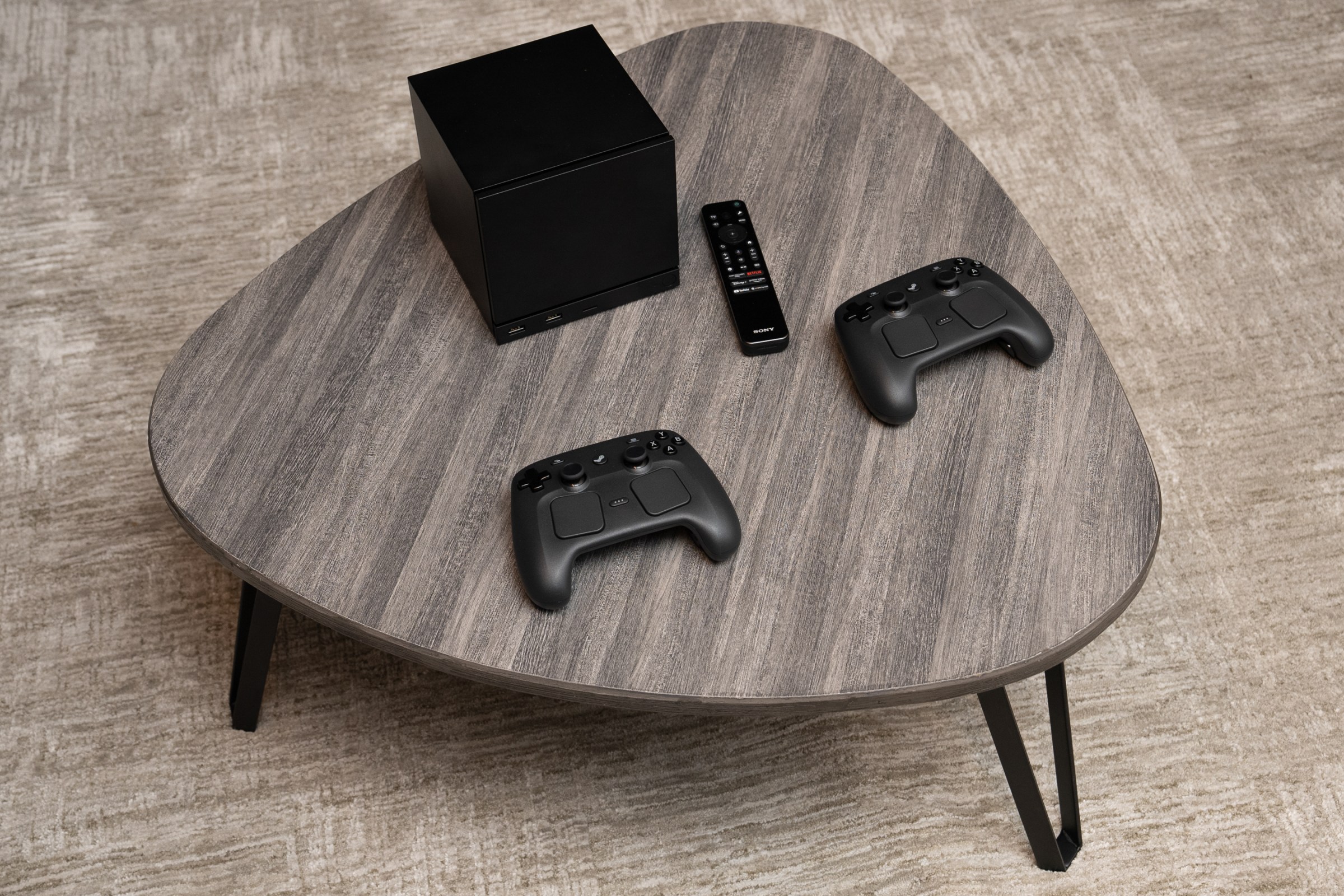 The black Steam Machine cube, next to a TV remote and two Steam Controllers, sitting on a rounded triangle of a small coffee table with a grey wood veneer finish.