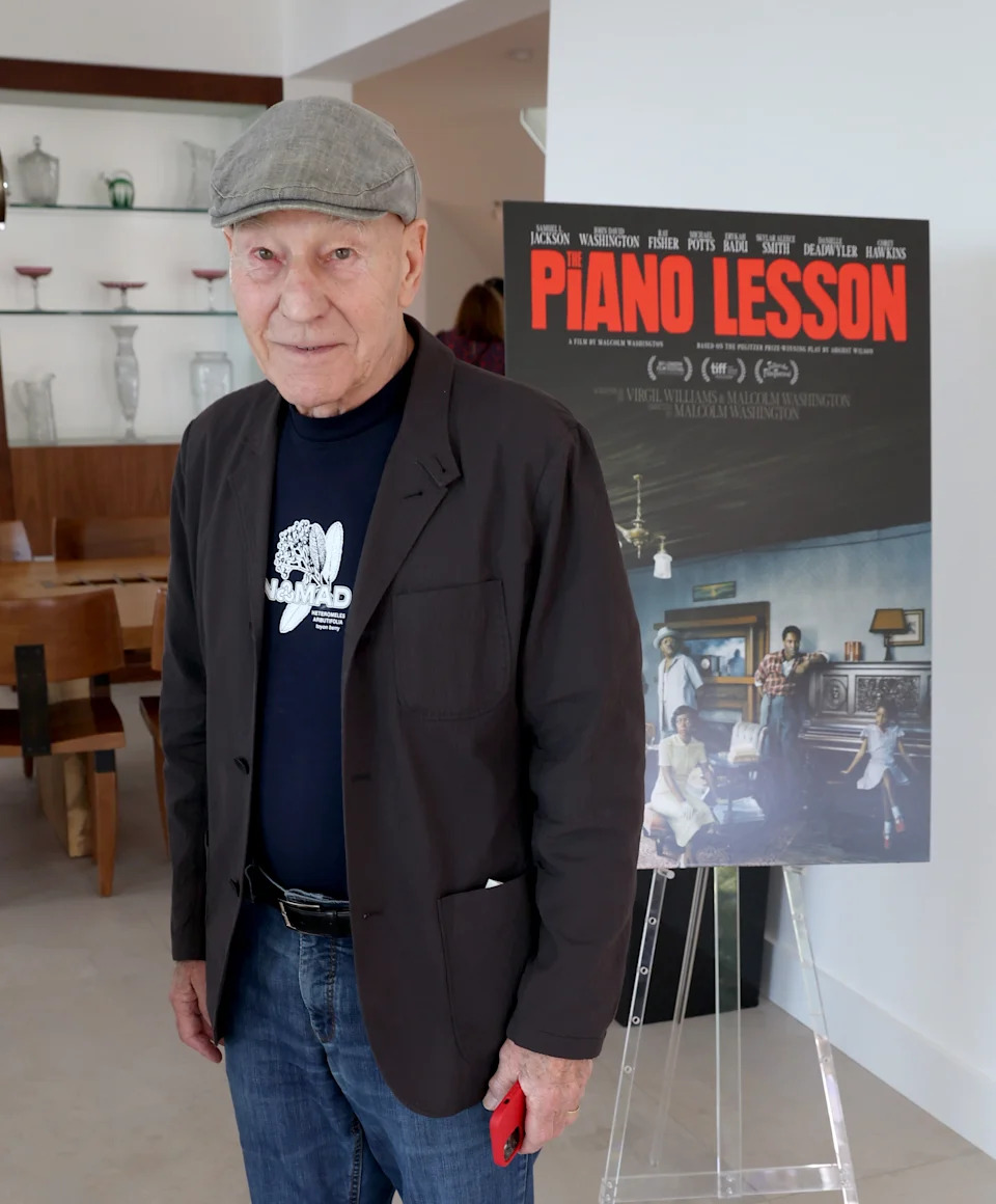Patrick Stewart wearing a flat cap and casual jacket stands beside a "Piano Lesson" movie poster