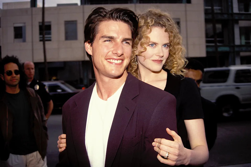 Two celebrities standing side by side, one wearing a suit jacket and T-shirt, the other in a casual top with curly hair, smiling outdoors