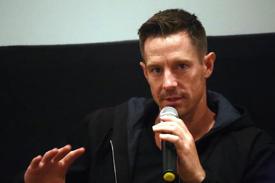 A person speaking into a microphone at a speaking event, gesturing with their hand, wearing a casual outfit with a hoodie