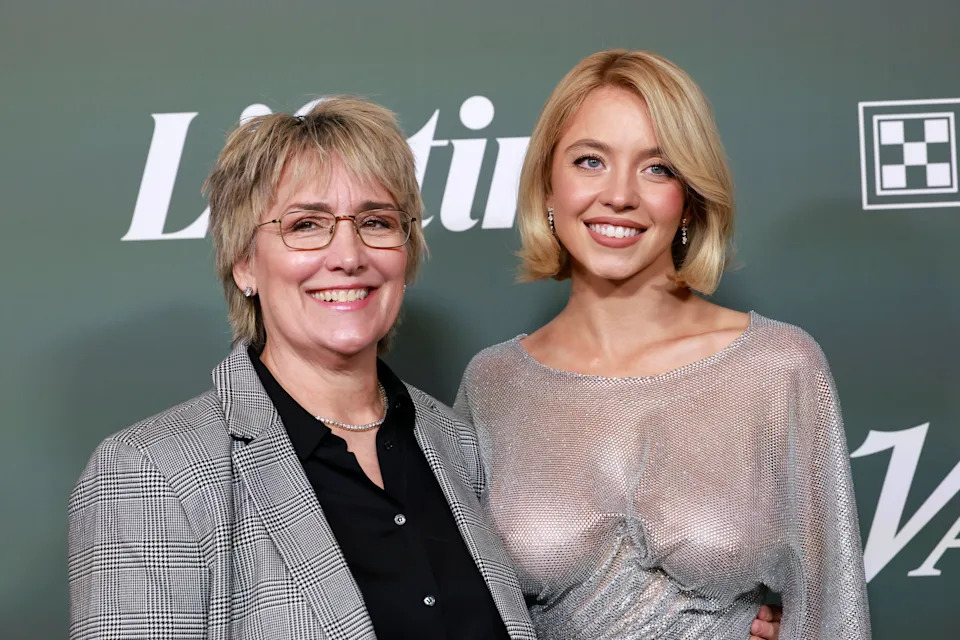 Two individuals pose on a red carpet; one wears a plaid blazer, the other a sheer, shimmering dress
