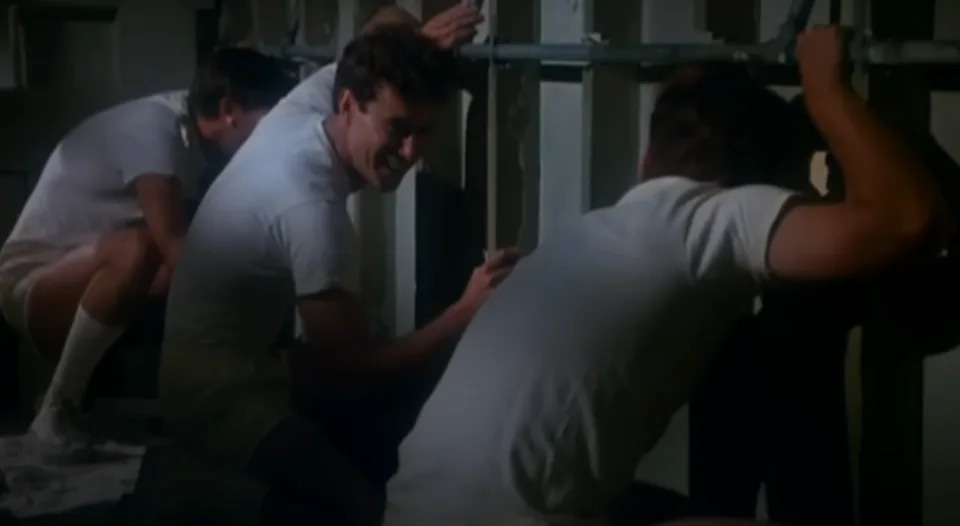 Three men in casual T-shirts and shorts work look through holes in a wall.