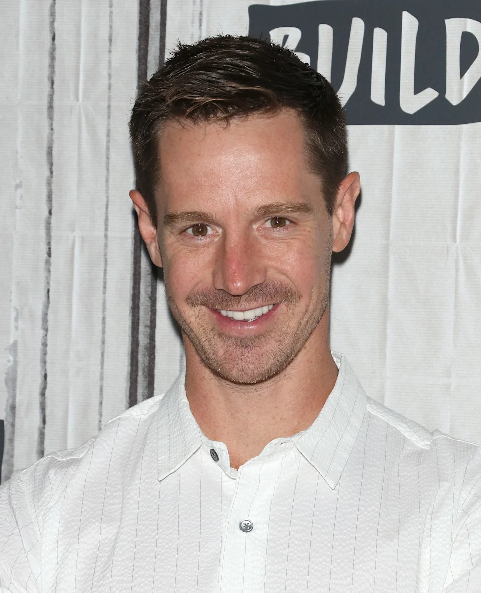 A person in a white textured shirt smiles at an event backdrop