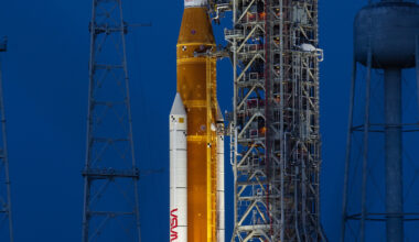 A large orange NASA rocket with white booster rockets stands vertically on a launch pad, lit by spotlights, with the full Moon and night sky behind it.