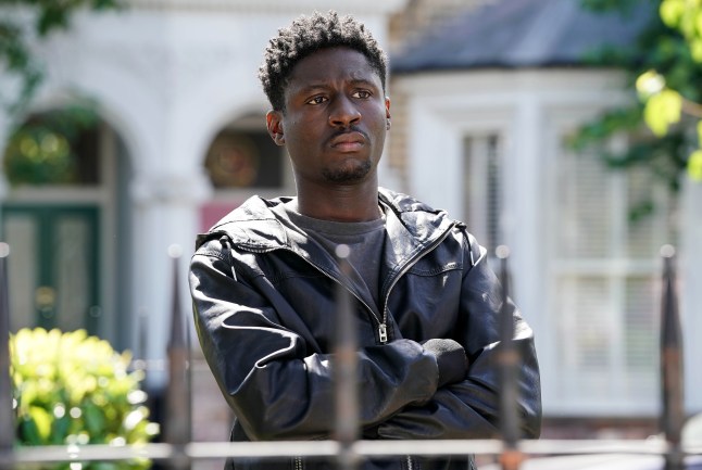 Okie standing in Albert Square in EastEnders, looking contemplative with his arms folded.