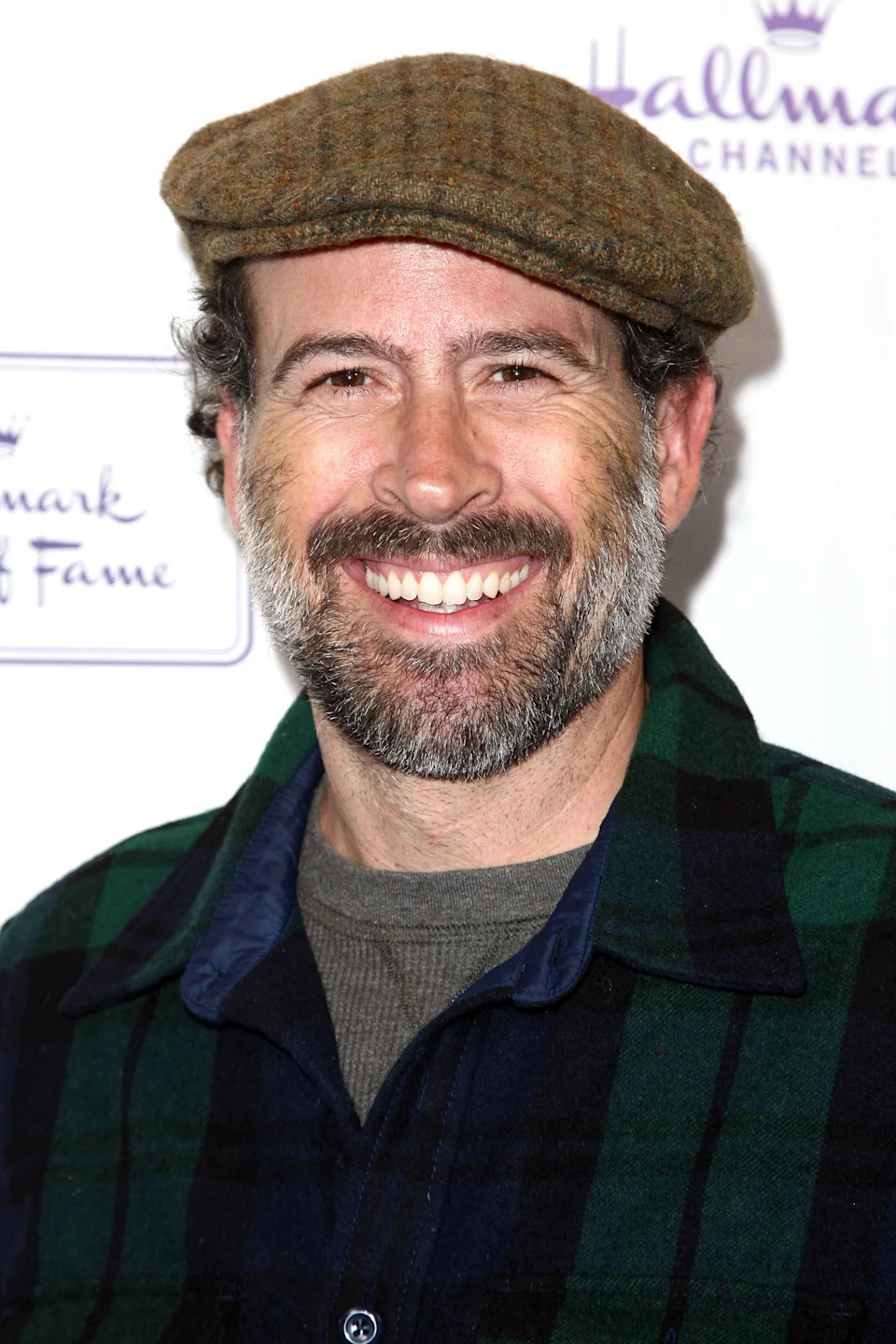 A smiling man with a beard wears a flat cap and plaid shirt at a Hallmark Channel event backdrop