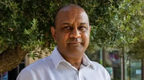 Getty Images Sanjay Shah standing outside underneath trees in what appears to be a garden in a built up office area - he is a middle aged man, with a white shirt and thinning hair. He is looking just past the camera with a neutral expression. 