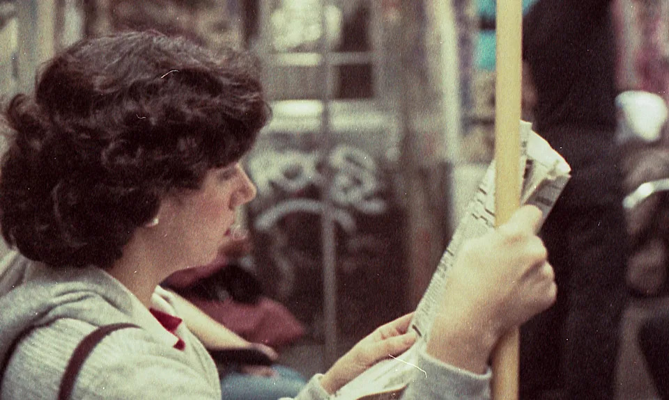 Person reading a newspaper on a graffiti-covered subway, gripping a yellow pole. Others are visible in the background