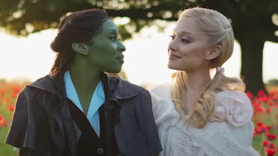 Cynthia Erivo's Elphaba and Ariana Grande's Glinda looking at each other smiling on the set of Wicked: For Good with poppy fields behind them.