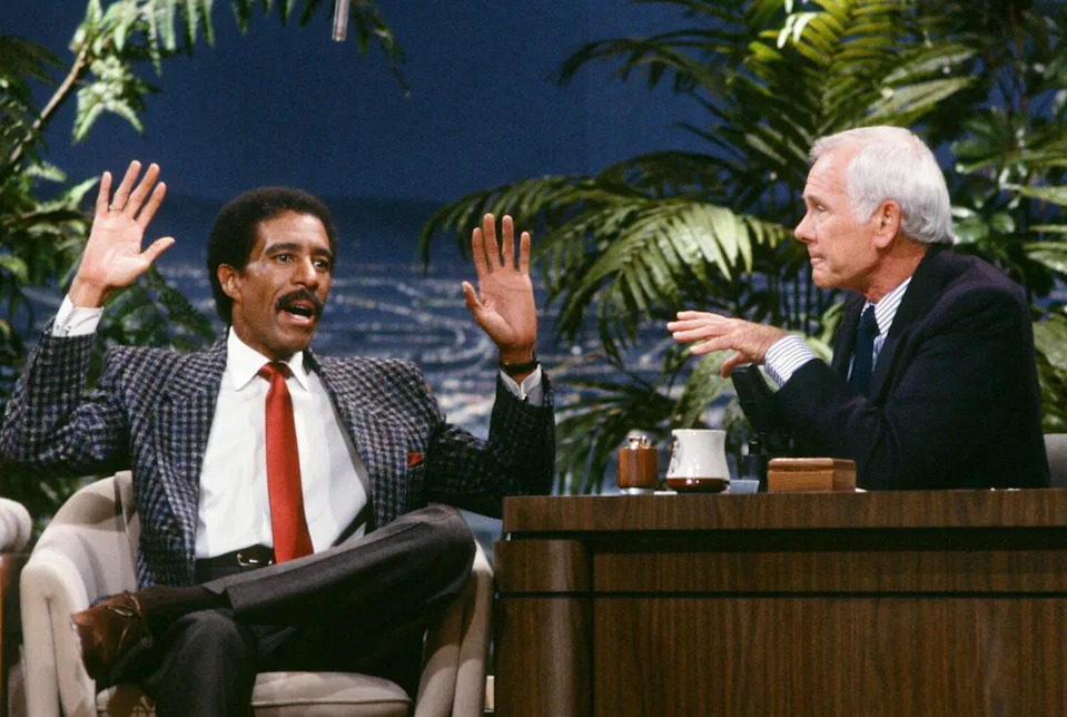 A man in a plaid jacket and red tie holds up his hands as he sits next to a man with white hair in a suit.