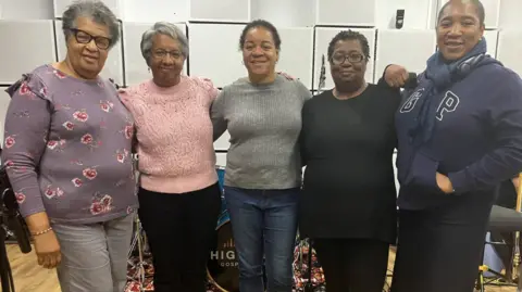 BBC From left Beverley Hepburn-Henderson, Kathy Hepburn-Hinds, Evadne Anderson, Avril Bartholomew and Angela Brown-Johnson all stand with their arms around one another. Beverley has a purple jumper on and grey trousers and glasses. Kathy is wearing a pink jumper and black trousers and has glasses on. Evadne is in a grey top and jeans. Avril is wearing a black top and trousers and wears glasses and Angela has a blue hoodie on with blue scarf and dark trousers.

