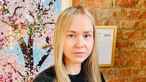 PSNI Iwona Giza. She has long blonde hair, wearing a black t-shirt. She is standing in front of a brick wall with a frame and a large floral picture on it.