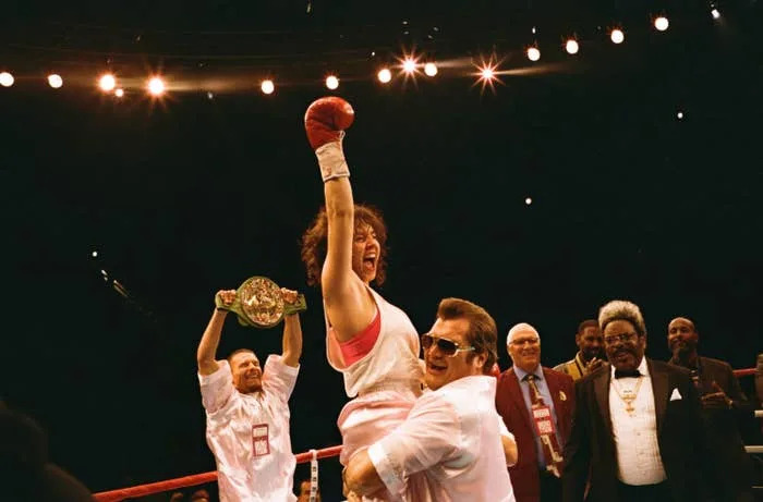 A victorious boxer celebrates in the ring, arm raised, as supporters and officials cheer, holding a championship belt nearby