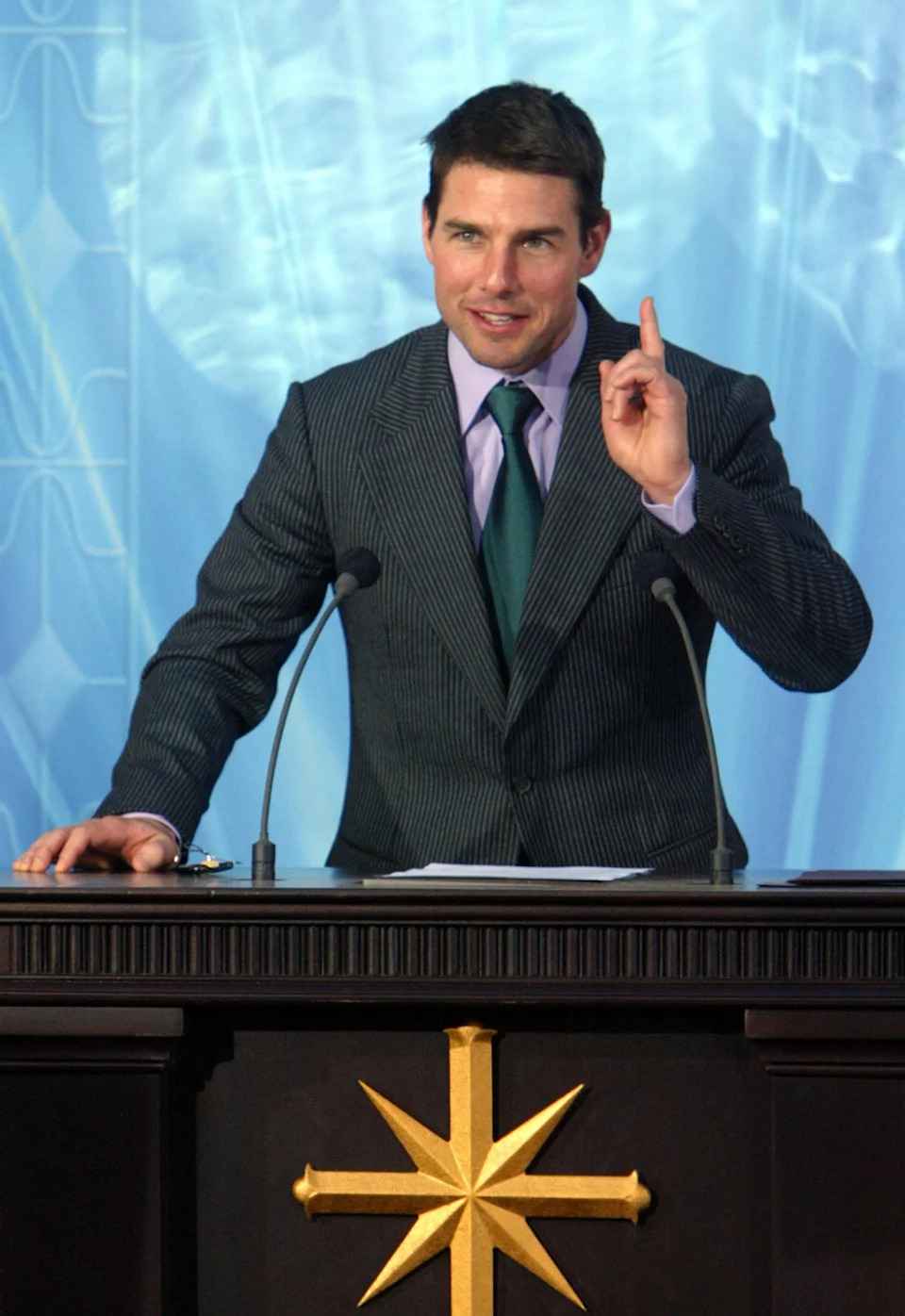 A person stands confidently at a podium, wearing a pinstripe suit and tie, while making a point with an upraised finger gesture