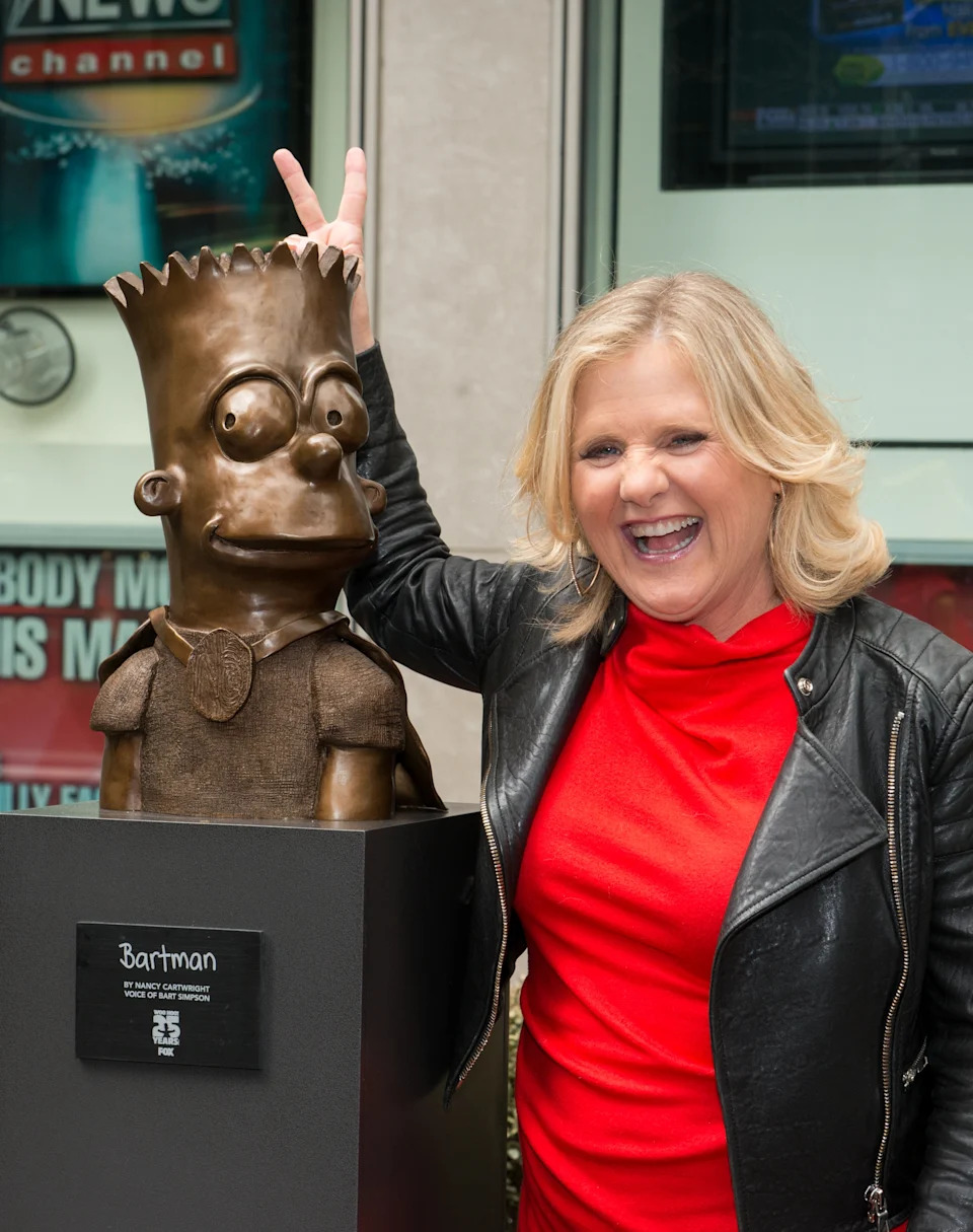 A woman playfully poses with a bronze statue of Bart Simpson, smiling and making a peace sign above its head