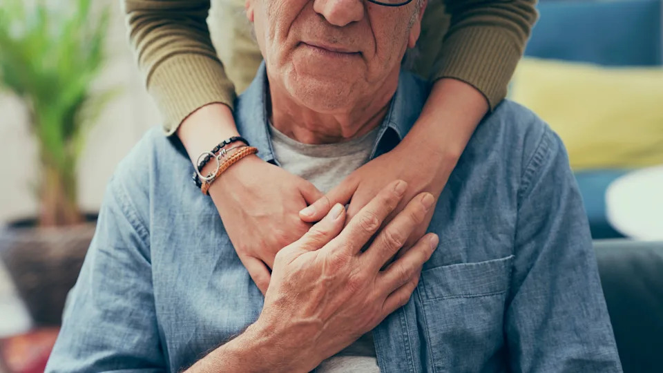 An elderly man in a denim shirt is warmly embraced from behind by another person, whose arms are wrapped around his shoulders