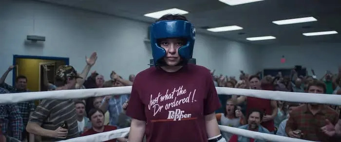 Person wearing headgear stands in a boxing ring, surrounded by cheering crowd, wearing a t-shirt with text about Dr Pepper
