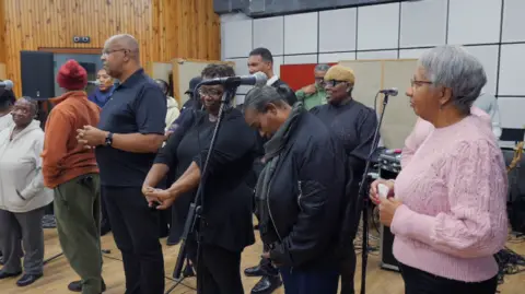 Men and women stand by microphones in the studio. They are wearing tops and trousers and some have coats on and are wearing glasses.