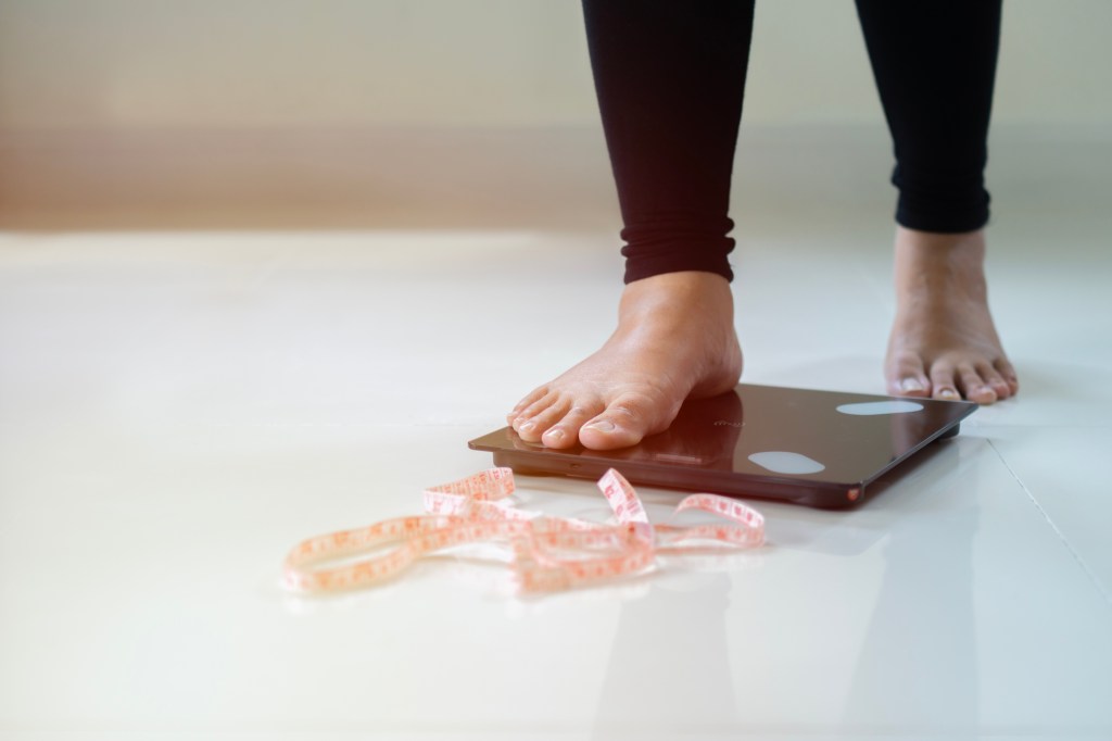 A person steps onto a dark bathroom scale with a tape measure on the floor beside it.