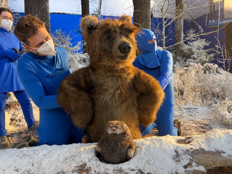 Three people in blue suits operate a realistic bear puppet and a small animal puppet on a snowy set with trees. Two puppeteers wear masks, and the background includes a blue screen for visual effects.