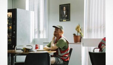 A man in a reflective vest sits at a table with snacks and a can of Coke, appearing thoughtful. Sunlight filters through vertical blinds. A vending machine, potted plant, and a framed portrait are in the background.